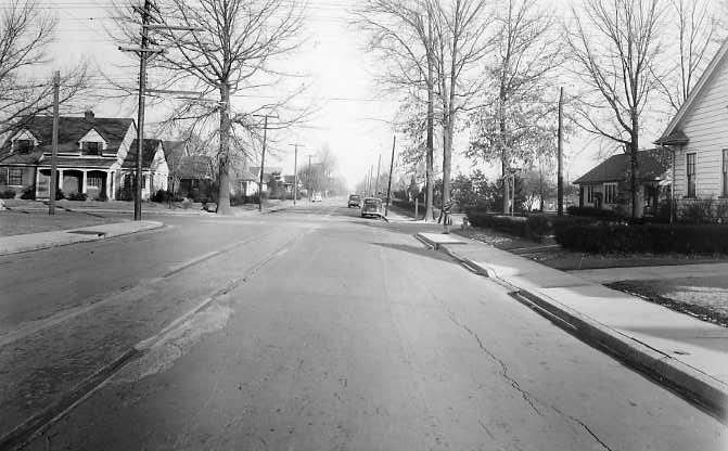 A black and white photo of a quiet residential street with houses, trees, and parked cars.