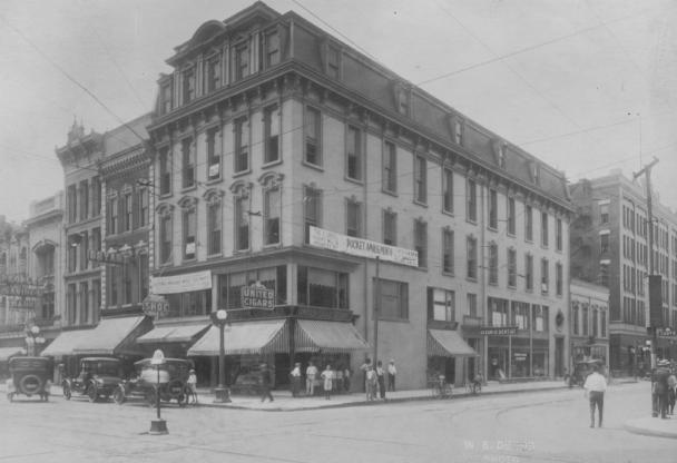 Vintage street corner with classic buildings, cars, and pedestrians.