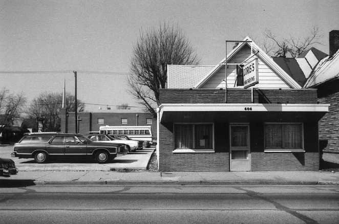 Vintage photo of a small building with "Dresses" sign and old cars parked alongside.