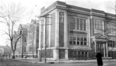 Historic building on a street corner with bare trees and a solitary figure walking by. Black & white photo.