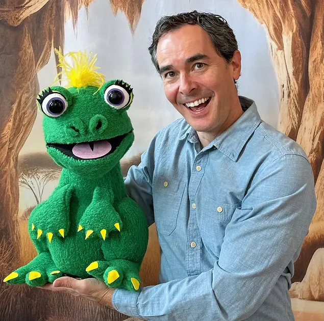 A man is smiling and holding a bright green dinosaur puppet with big eyes and a playful expression against a scenic backdrop.