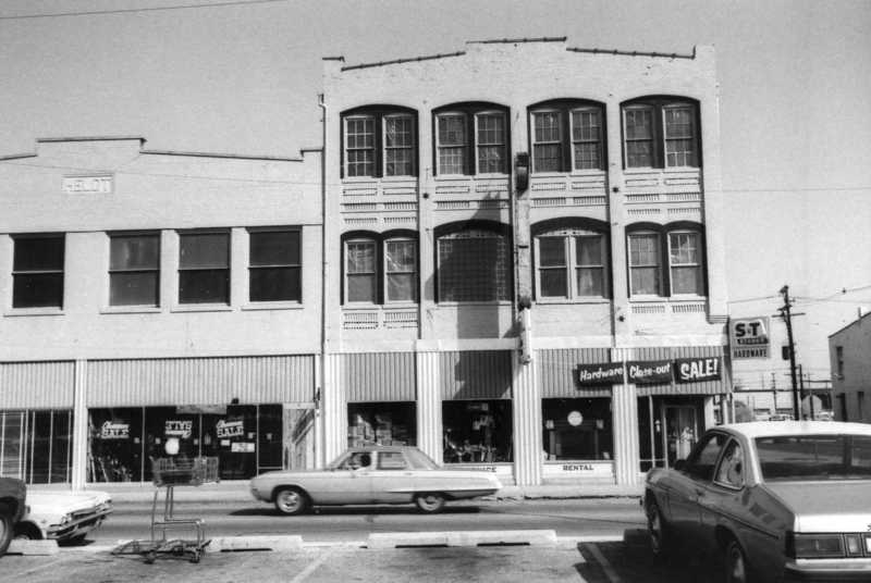 Vintage street view with cars, a two-story building labeled "Hardware," and a clear-out sale sign. Black and white photo.