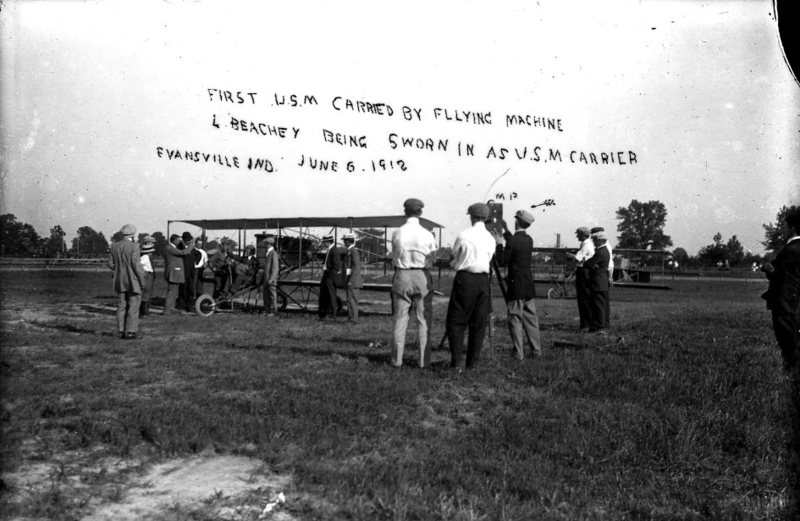 Historic photo of people at an airfield with a biplane and a banner about the first U.S. mail carried by flying machine, dated June 6, 1912.