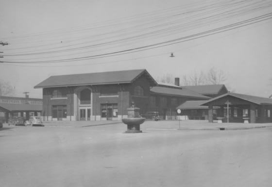 Vintage train station with tracks, buildings, and a fountain, in grayscale.