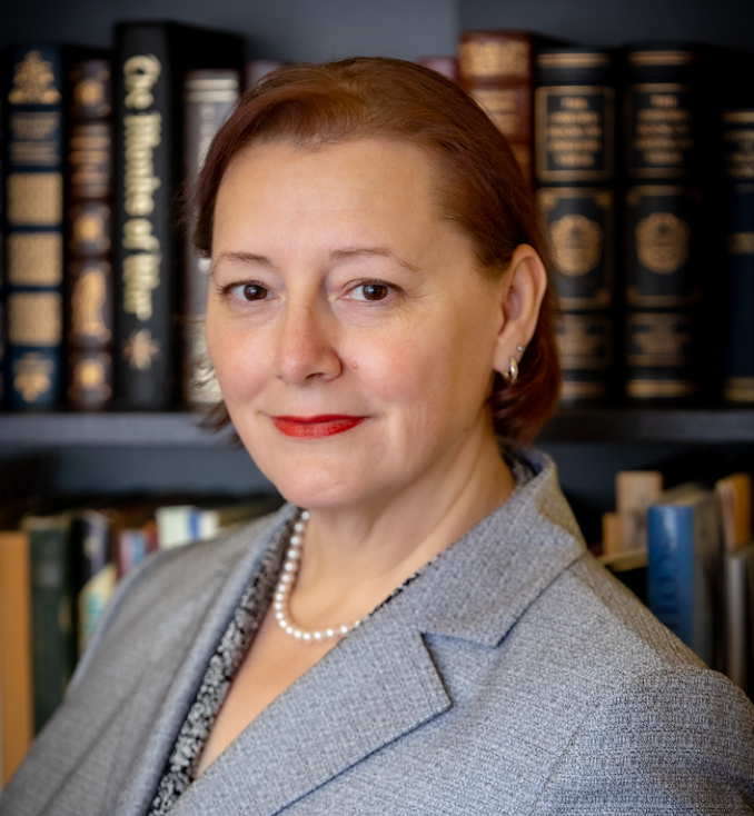 A person in a suit with a pearl necklace stands in front of a bookshelf filled with various books.