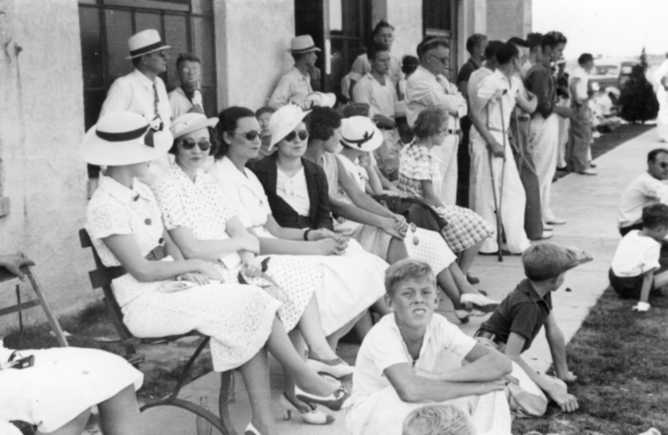 Group of vintage-dressed people sitting & standing, possibly at a social event.