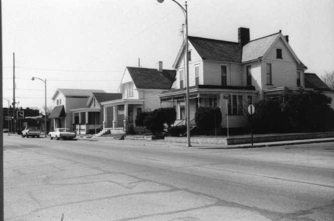 Black and white photo of a street with traditional houses and parked cars.