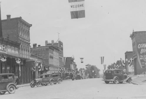 Historic street lined with old buildings and vintage cars, with a "Welcome" banner overhead.