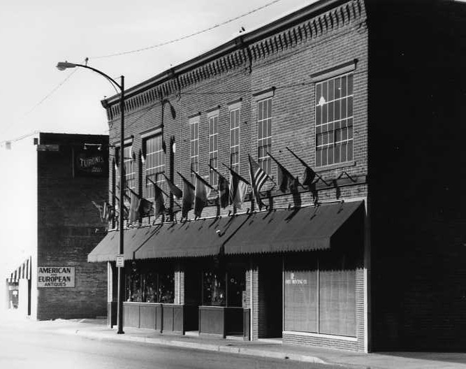 A black and white photo of a building with multiple American flags and a ghost sign reading "AMERICAN EUROPEAN".