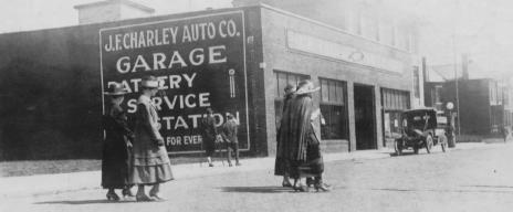 Vintage street scene with a garage, people walking, and an old car.