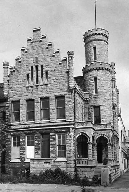 An old castle-like building with a turret, crenellations, and arched entrance. Black and white photo.