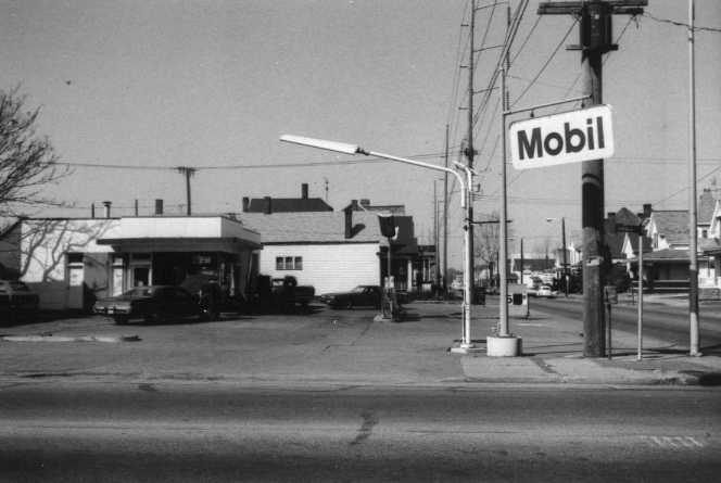 Black and white photo of a street scene with a Mobil gas station sign and a building behind it.