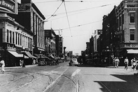 Vintage street scene with tramlines, old cars, and pedestrians, likely in early 20th century.