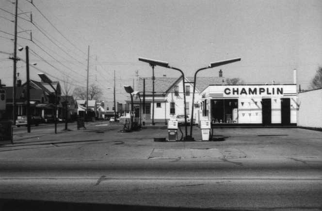 Black & white photo of an old gas station with "CHAMPLIN" sign, fuel pumps in the front, and other buildings in the background.
