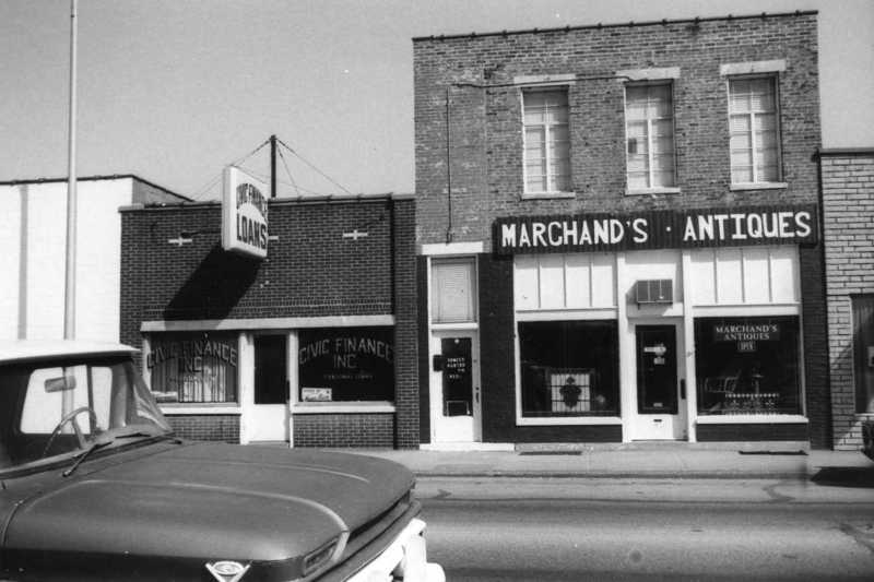 Black and white photo of a vintage car in front of buildings with signs saying "Loans" and "Marchand's Antiques".