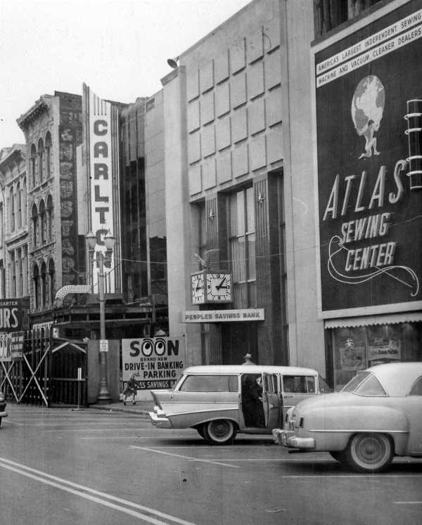 Vintage street scene with old cars, Carlton theater sign, Atlas Sewing Center ad, and "People's Savings Bank" signage.