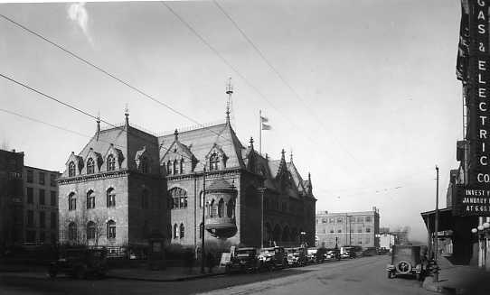 Vintage street scene with ornate building, cars parked alongside, and electric advertisements on the right.