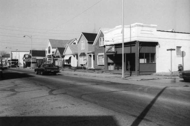 Black and white photo of a street with cars, houses, and a storefront.