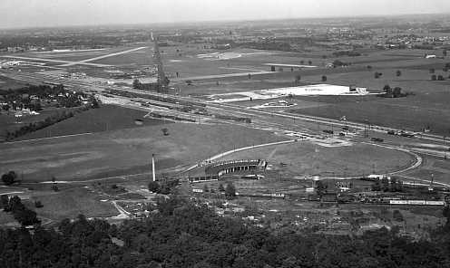 Aerial view of a landscape with fields, roads, and structures, possibly an airport or racetrack. Black and white photo.