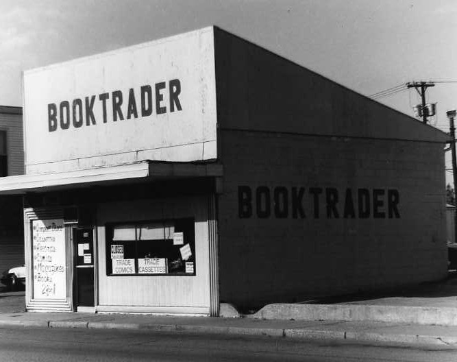 A black and white photo of a corner bookstore named "BOOKTRADER" with signs on the windows.