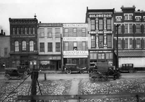 Old city street with vintage cars and buildings with signs for "Furniture House," "Pleasing Supplies," and "Carriage Time."