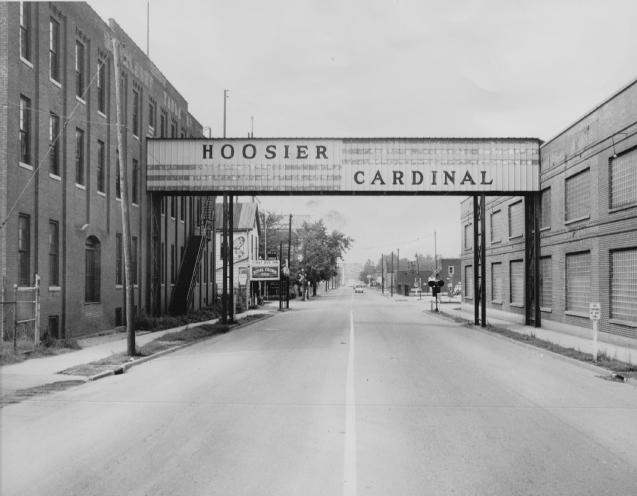 An old black and white photo of a street with an overhead sign reading "HOOSIER CARDINAL" between two buildings.