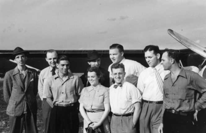A group of people, possibly from the mid-20th century, standing in front of an airplane, dressed in period attire.