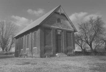 A black and white photo of an old, small, brick schoolhouse with a pitched roof under a cloudy sky.
