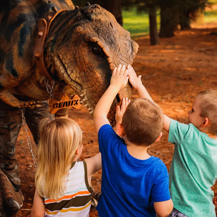 Three kids are happily touching the face of a life-like dinosaur costume in a natural setting.