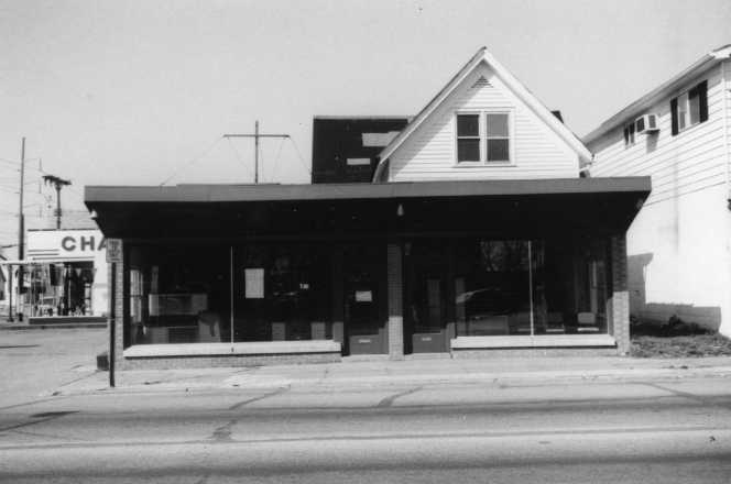 A black-and-white photo of a street view with a shopfront and houses.