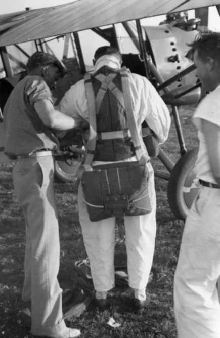 Three individuals by an aircraft, one wearing a parachute harness, likely preparing for a flight or jump.