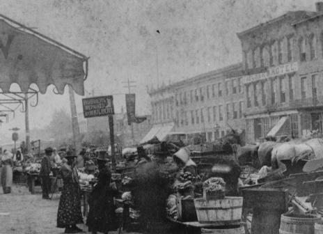 A historical black and white photo of a busy outdoor market with horses, carts, and people, in a town setting.