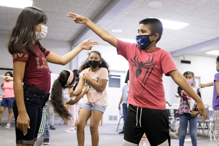 Children wearing masks engage in a playful interaction, likely during a game or activity in a classroom setting.