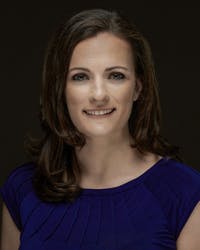 A smiling woman with brown hair in a blue top against a dark background.