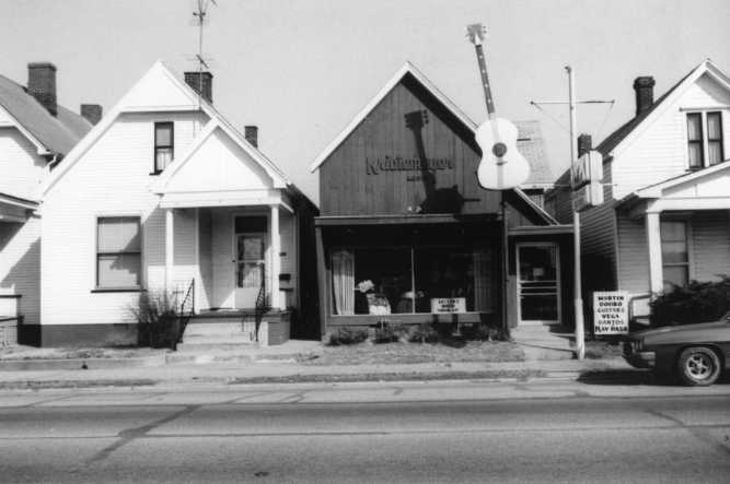 Black and white photo of a street with houses and a building with a large guitar sign, captioned "Kilgore's Music."