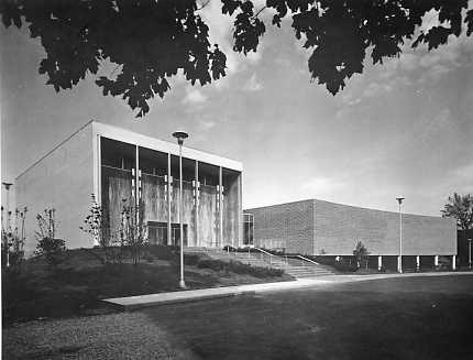 A black and white photo of a modern building with large windows and a clear sky, framed by tree foliage at the top.