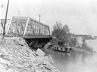 A black and white photo of a truss bridge over a river, with a boat underneath and construction equipment nearby.