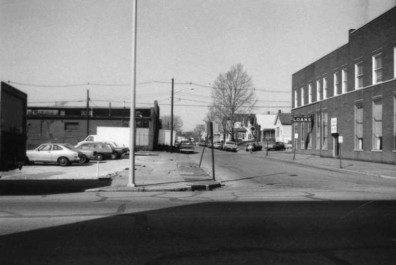 Black & white photo of a street scene with old cars, buildings, a "Loans" sign, and no visible people.
