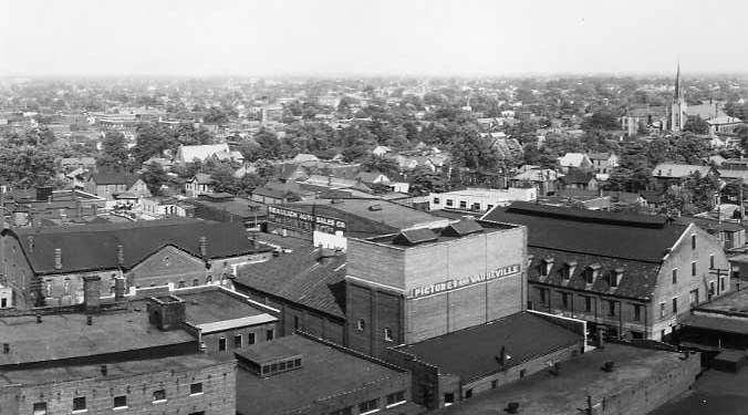Black and white aerial view of a vintage town with buildings, including a theater, and a church spire in the background.