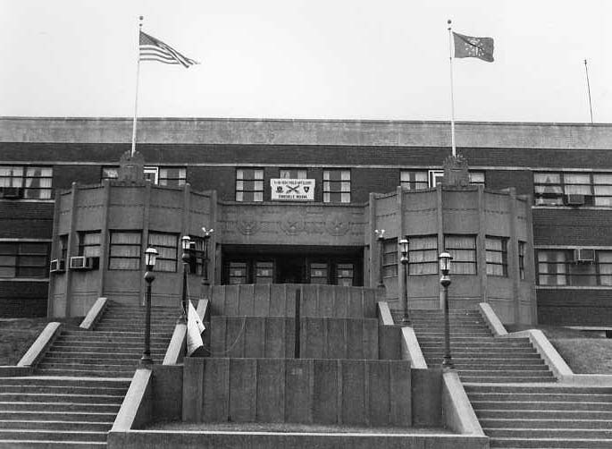 An old building with a flag on the roof and a crest-like sign above the entrance, flanked by two lamps and a staircase.
