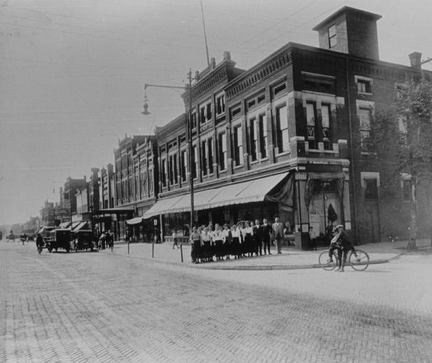 Historic street scene with brick buildings, horse-drawn carriages, and a cyclist on cobbled streets.