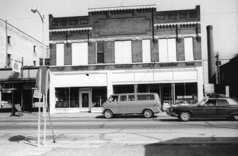 Old buildings with storefronts, a vintage van, a car, and a streetlight on an urban street.