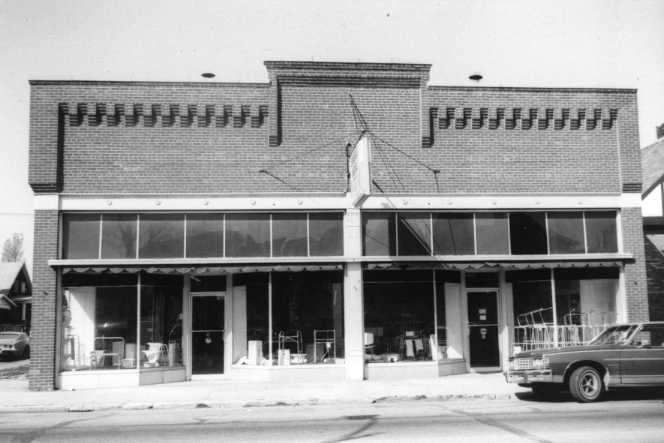 A black-and-white photo of a vintage two-story storefront with large display windows and a classic car parked in front.