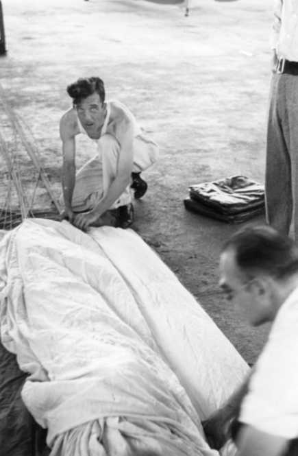 A black and white photo showing a man crouching, looking towards the camera, with others and a covered object nearby.