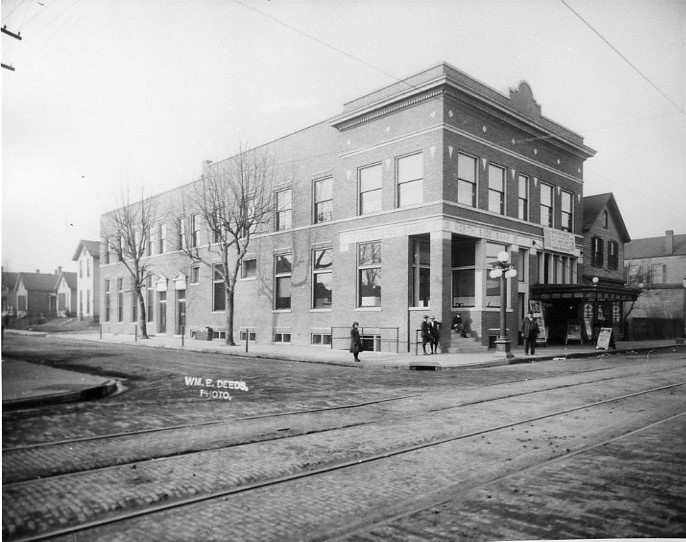 Black and white photo of an early 20th-century corner building with tram tracks, people, and bare trees.