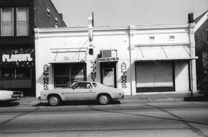 A black and white photo of a street scene with an adult bookstore and a car parked in front.