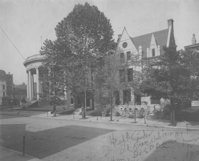 Historic black and white photo of a street corner with classical-style buildings and trees.