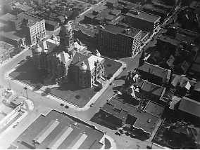 Black and white aerial view of an urban area with buildings, streets, and shadows indicating bright sunlight.