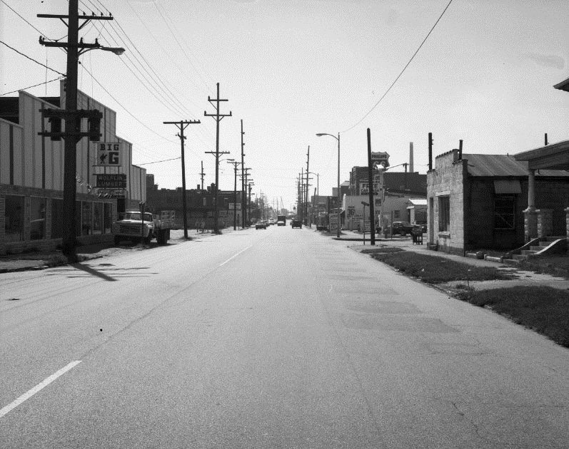 Empty street in a town with power lines, storefronts, and parked cars. Appears to be an old, black and white photo.