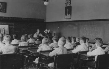 A black and white photo of students in an old-fashioned classroom, focusing on the teacher.